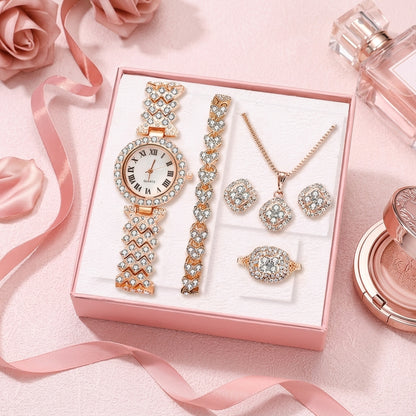A women's jewelry set displayed in a pink gift box, featuring a watch with a crystal-embellished band and round face, a crystal bracelet, a crystal necklace, matching crystal earrings, and a crystal ring. The set is styled on a pink background with roses and perfume bottles.