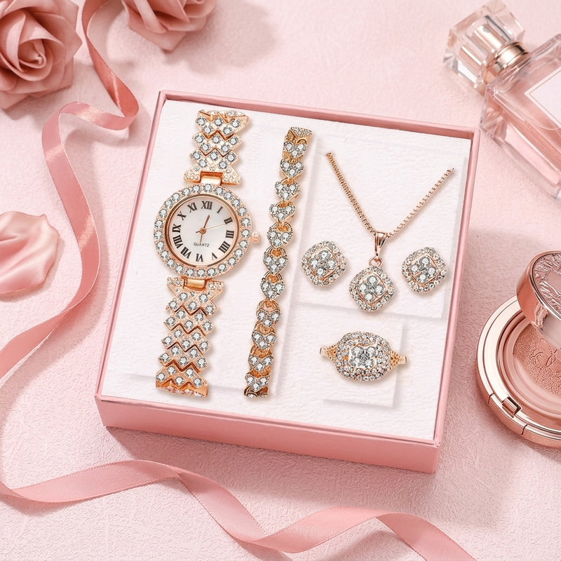 A women's jewelry set displayed in a pink gift box, featuring a watch with a crystal-embellished band and round face, a crystal bracelet, a crystal necklace, matching crystal earrings, and a crystal ring. The set is styled on a pink background with roses and perfume bottles.
